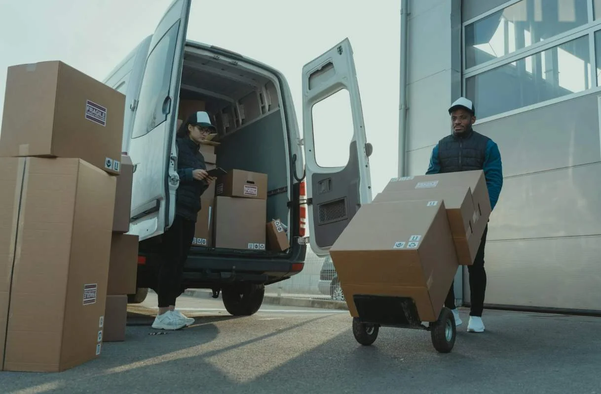 homme poussant un chariot avec des colis devant un camion de livraison avec des colis. Il assure le bon ordonnancement de la logistique.